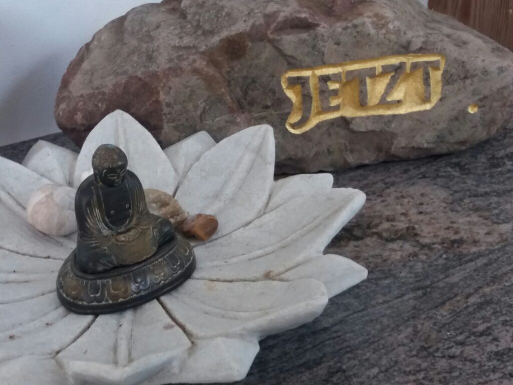 Zen-Statue auf Lotusblüte vor einem Stein mit der Aufschrift „Jetzt“ – Symbol für Achtsamkeit und innere Einkehr im Jahreskreis